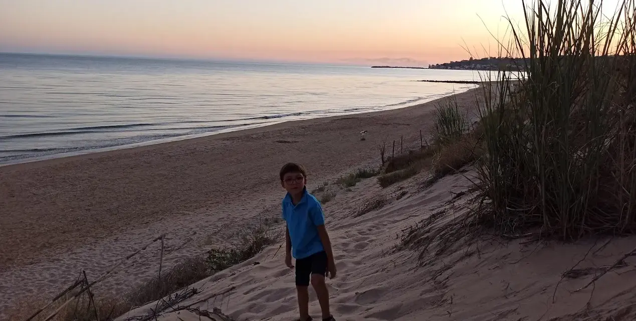 Bambino con occhiali su una duna di sabbia a Porto Palo, con il mare calmo e il cielo al tramonto sullo sfondo.