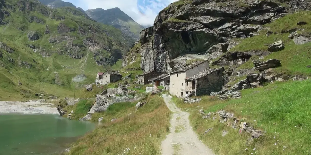 Antico borgo in pietra nel **Val di Susa**, con un sentiero che sale verso le case arroccate su un pendio verdeggiante, vicino a un lago alpino.