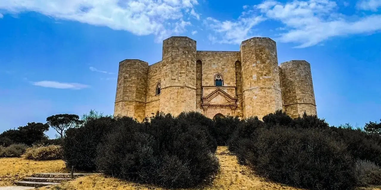 Castel del Monte, un'icona pugliese, si erge maestoso su una collina, circondato da una vegetazione rigogliosa sotto un cielo azzurro.