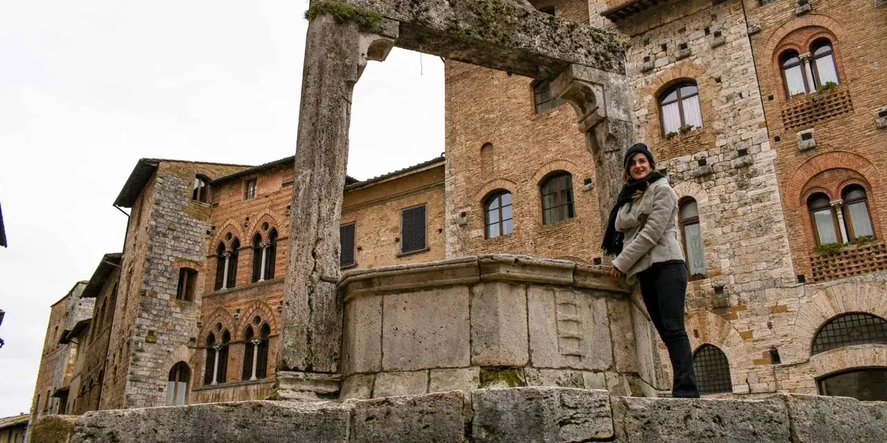 Donna sorride vicino all'arco in pietra e al pozzo di San Gimignano, con palazzi medievali sullo sfondo.