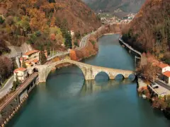 Il **ponte della Maddalena**, con i suoi archi irregolari, attraversa un fiume circondato da colline autunnali e case colorate.