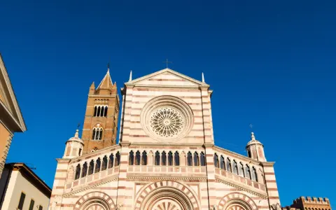 Il Duomo di Grosseto, con la sua facciata a strisce bianche e rosa e il rosone centrale, si staglia contro un cielo azzurro intenso.