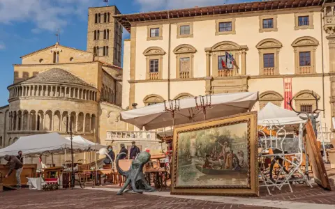 Mercatino d'antiquariato in piazza Grande ad Arezzo, con opere d'arte, sculture e mobili esposti sotto il sole.