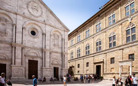 Piazza Pio II a Pienza, con la facciata del Duomo e il Palazzo Piccolomini. Turisti passeggiano nella piazza soleggiata.