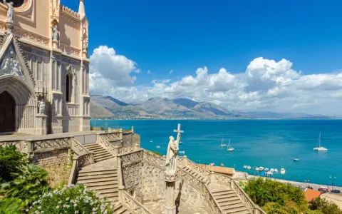 La chiesa domina il mare azzurro della Riviera di Ulisse. Scale imponenti conducono a un portale gotico, mentre barche solcano le acque.