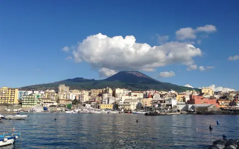 Vista del porto di Torre del Greco con il Vesuvio sullo sfondo, sotto un cielo azzurro con nuvole bianche.