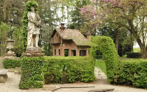 Giardino incantato a Grazzano Visconti con statua romana, casetta in mattoni e sentiero tra siepi fiorite.