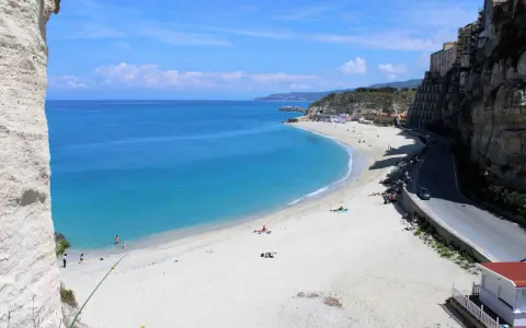 Spiaggia bianca e mare cristallino sulla **Costa degli Dei**. Case aggrappate alla scogliera, una strada panoramica e bagnanti che si godono il sole.