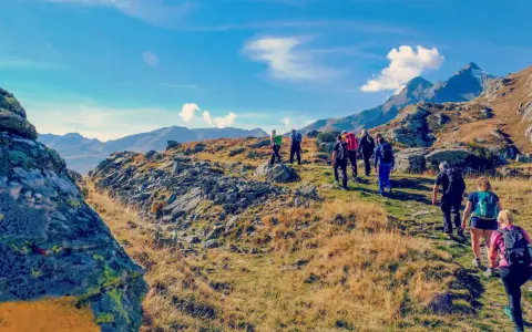 Un gruppo di escursionisti cammina su un sentiero roccioso e erboso, con montagne sullo sfondo. Il **Passo dello Spluga** offre panorami mozzafiato.