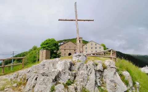 Croce di legno a San Pellegrino in Alpe, con edifici storici sullo sfondo e rocce in primo piano.