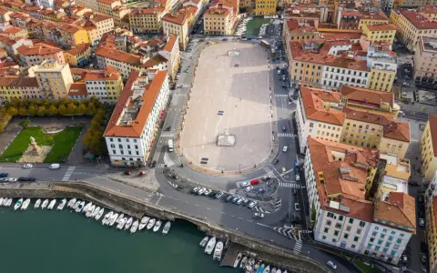 Vista aerea della **Piazza della Repubblica a Livorno**, con edifici storici, un monumento centrale, auto e barche ormeggiate nel canale.