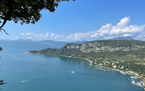 Vista panoramica del Lago di Garda con San Zeno di Montagna sullo sfondo, tra montagne verdi e cielo azzurro.