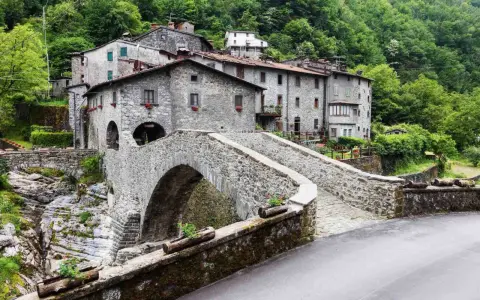 Ponte in pietra a Vergemoli, con case in pietra e vegetazione lussureggiante sullo sfondo. Le antiche fabbriche di Vergemoli sembrano emergere dalla collina.