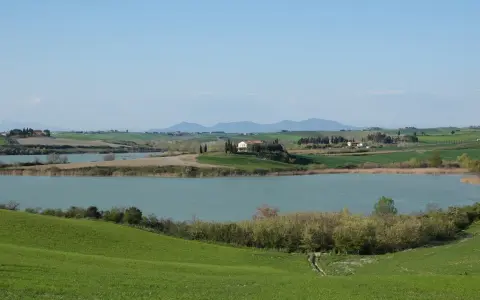 Campagna toscana con lago e casale in collina, un luogo dove la **santa luce cosa vedere** è ovunque, tra dolci pendii verdi e il cielo azzurro.