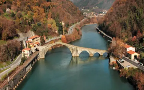 Il **ponte della Maddalena**, con i suoi archi irregolari, attraversa un fiume circondato da colline autunnali e case colorate.