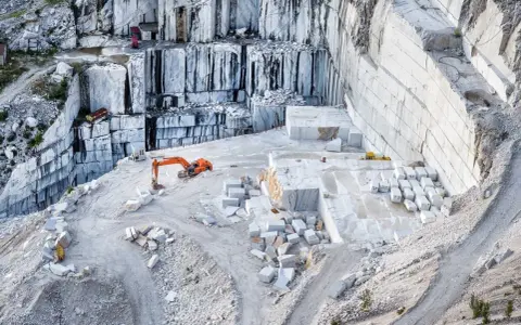 Cava di marmo a Carrara, con escavatore arancione al lavoro tra blocchi di pietra bianca. Un luogo affascinante da vedere a Carrara.