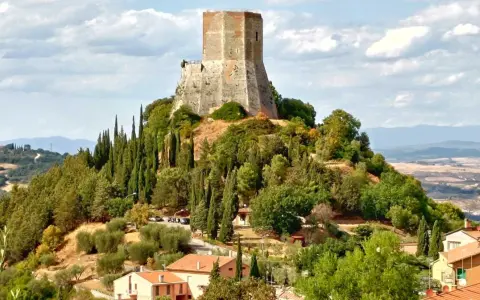 La rocca di Tentennano domina la collina, circondata da cipressi e ulivi. Sotto, un borgo con tetti rossi si adagia nel paesaggio toscano.