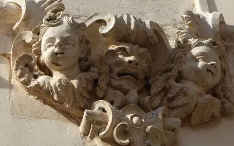 Dettaglio scultoreo barocco a Martina Franca: volti di putti e una maschera grottesca emergono dalla pietra calcarea, testimoni di un'antica arte.