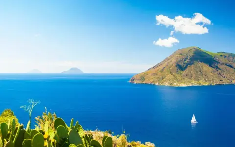 Panoramica delle Isole Eolie: cosa vedere? Un veliero solca il blu intenso del mare, con isole vulcaniche sullo sfondo e fichi d'India in primo piano.