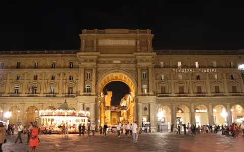 La piazza della Repubblica a Firenze di notte, con il suo arco monumentale illuminato, gente che passeggia e una giostra scintillante.