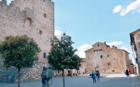 Piazza nel centro storico di Castellina in Chianti con torre medievale, edifici in pietra e scultura.