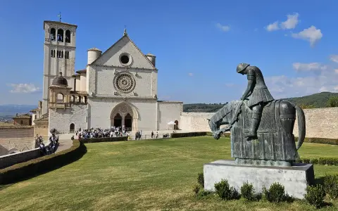 Statua di un cavaliere pensieroso davanti alla maestosa basilica San Francesco d'Assisi, sotto un cielo azzurro.