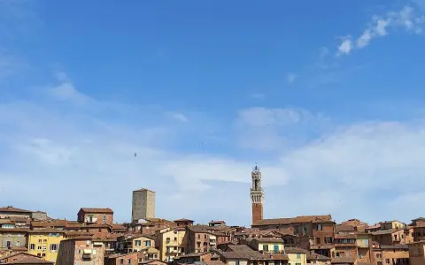 Panoramica di Siena con la Torre del Mangia che svetta tra i tetti rossi, sotto un cielo azzurro con nuvole bianche.