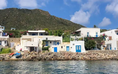 Case bianche con porte blu sull'isola di Panarea, con il mare calmo e una collina verde sullo sfondo.
