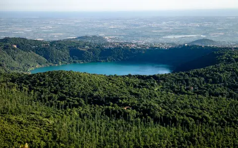 Vista panoramica del lago di Albano, circondato da rigogliosa vegetazione, con il borgo di Rocca di Papa in lontananza.