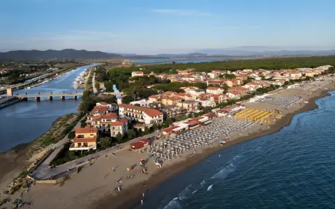Vista aerea di Castiglione della Pescaia: un canale navigabile con barche, case con tetti rossi e una spiaggia attrezzata con ombrelloni e lettini, bagnata dal mare.