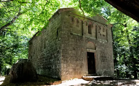Antica pieve in pietra nel bosco Isabella, Radicofani. La luce filtra tra gli alberi illuminando la facciata.