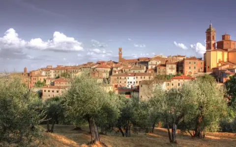 Il centro storico di Sinalunga si staglia su una collina, circondato da ulivi secolari sotto un cielo azzurro con nuvole bianche.