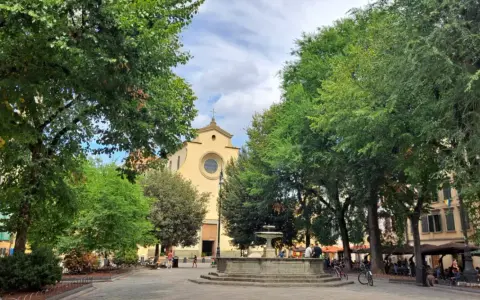 Piazza Santo Spirito a Firenze, con la chiesa sullo sfondo, una fontana al centro e alberi rigogliosi che incorniciano la scena.