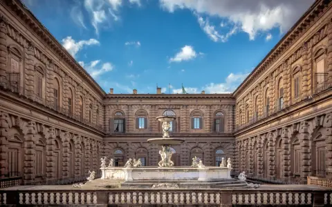 Cortile del Palazzo Pitti con fontana e statue, sotto un cielo azzurro con nuvole bianche.