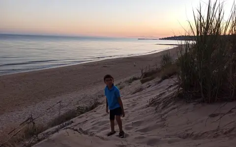 Bambino con occhiali su una duna di sabbia a Porto Palo, con il mare calmo e il cielo al tramonto sullo sfondo.