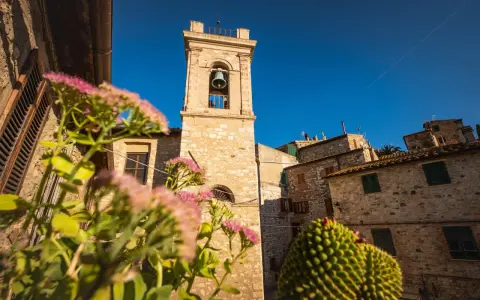 Campanile in pietra in un borgo delle Colline Metallifere, con fiori rosa in primo piano e cielo azzurro.
