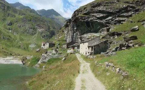 Antico borgo in pietra nel **Val di Susa**, con un sentiero che sale verso le case arroccate su un pendio verdeggiante, vicino a un lago alpino.