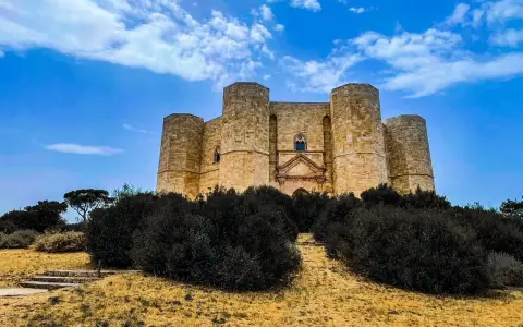 Castel del Monte, un'icona pugliese, si erge maestoso su una collina, circondato da una vegetazione rigogliosa sotto un cielo azzurro.