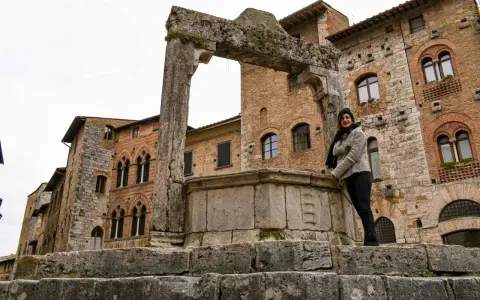 Donna sorride vicino all'arco in pietra e al pozzo di San Gimignano, con palazzi medievali sullo sfondo.
