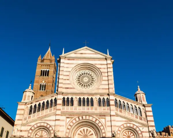 Il Duomo di Grosseto, con la sua facciata a strisce bianche e rosa e il rosone centrale, si staglia contro un cielo azzurro intenso.