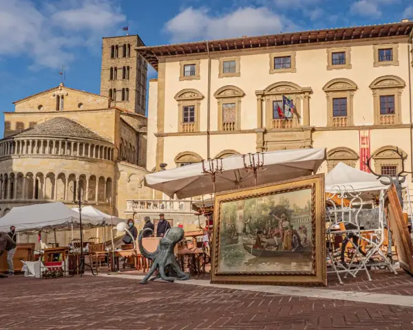 Mercatino d'antiquariato in piazza Grande ad Arezzo, con opere d'arte, sculture e mobili esposti sotto il sole.