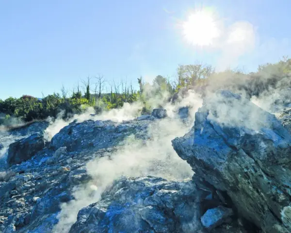 Fumarole attive nella valle del diavolo, con rocce grigie e blu avvolte dal vapore. Il sole splende nel cielo azzurro.