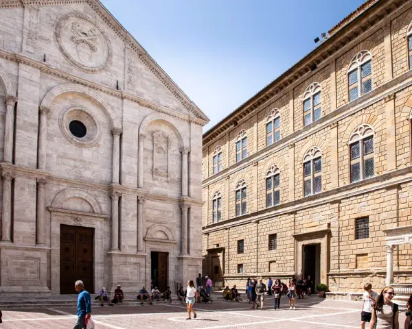 Piazza Pio II a Pienza, con la facciata del Duomo e il Palazzo Piccolomini. Turisti passeggiano nella piazza soleggiata.