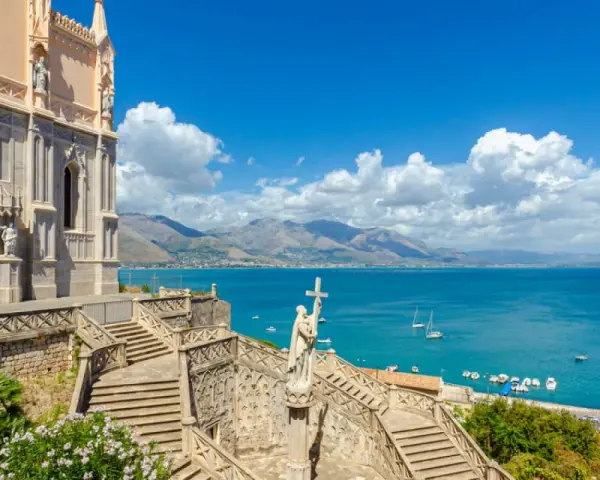 La chiesa domina il mare azzurro della Riviera di Ulisse. Scale imponenti conducono a un portale gotico, mentre barche solcano le acque.