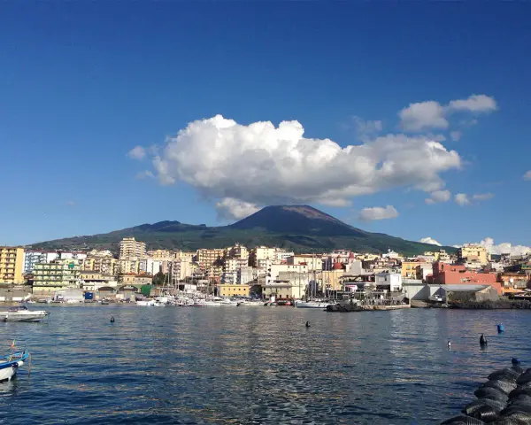 Vista del porto di Torre del Greco con il Vesuvio sullo sfondo, sotto un cielo azzurro con nuvole bianche.