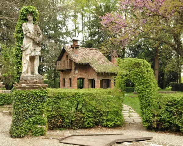 Giardino incantato a Grazzano Visconti con statua romana, casetta in mattoni e sentiero tra siepi fiorite.