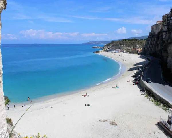 Spiaggia bianca e mare cristallino sulla **Costa degli Dei**. Case aggrappate alla scogliera, una strada panoramica e bagnanti che si godono il sole.