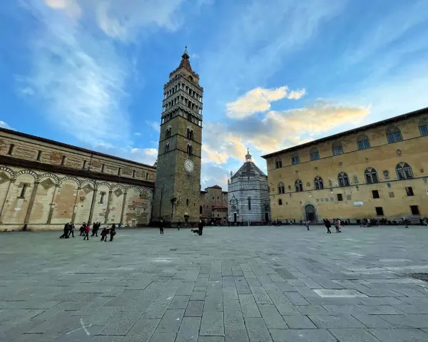Piazza del Duomo di Pistoia con la torre campanaria, il battistero e il palazzo vescovile sotto un cielo nuvoloso.