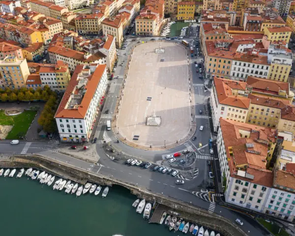 Vista aerea della **Piazza della Repubblica a Livorno**, con edifici storici, un monumento centrale, auto e barche ormeggiate nel canale.