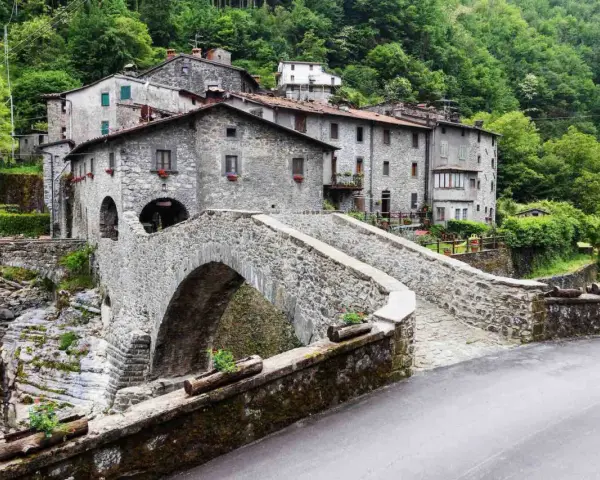 Ponte in pietra a Vergemoli, con case in pietra e vegetazione lussureggiante sullo sfondo. Le antiche fabbriche di Vergemoli sembrano emergere dalla collina.